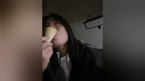 Video of an Asian woman with long black hair, wearing a dark jacket, eating a piece of cracker in a dimly lit room.