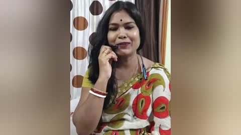 Video of an Indian woman in a colorful sari, holding a phone, with a background of a polka-dotted curtain and wooden door.