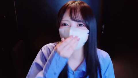 Video of a young Asian woman with long black hair and light skin, wearing a blue shirt and a white face mask, coughing into her hand against a dimly lit, dark background.