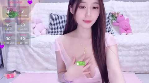 Video of an Asian woman with long brown hair, fair skin, and a slender physique, wearing a pink top, sitting on a pink mat in a cozy, white-walled room with a fluffy couch and teddy bears.