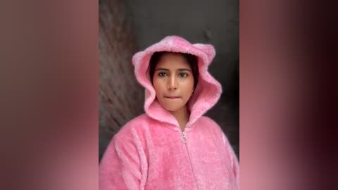 Video of a young woman with olive skin and dark hair, wearing a pink fluffy hooded robe, standing against a blurred, textured brick wall.