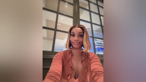 Video of a young woman with light brown skin and a short, straight bob hairstyle, wearing a coral pink ruffled blouse, smiling. Background features large glass windows with a modern building outside.