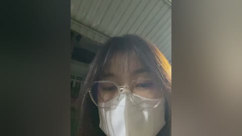 Video of a person with long, straight brown hair wearing clear glasses, a white face mask, and a black top. The background features a dimly lit, cluttered room with metal walls.