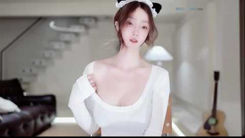 Video of a young East Asian woman with fair skin, wearing a white maid outfit with a black cat headband, standing in a modern, minimalist living room with a staircase and guitar in the background.