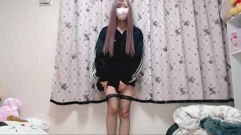 Video of an East Asian woman with long, straight, light brown hair, wearing a black Adidas tracksuit, fishnet stockings, and black lace garter belt, standing in front of a patterned white curtain, with a bed and stuffed toys in the background.