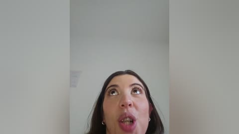 A close-up video of a woman with long dark hair, looking up with wide eyes and parted lips, against a plain, light-colored background.