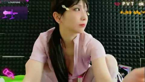 Video of a young Asian woman with long black hair and light skin, wearing a pink shirt, sitting on a bed with a black, grid-patterned wall behind her.