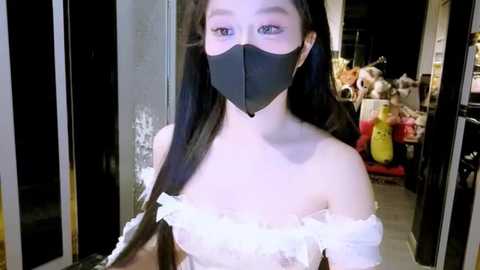 Video of an East Asian woman with long, straight black hair, wearing a black mask and a white, off-the-shoulder dress, standing in a dimly-lit, cluttered room with cardboard boxes and toys in the background.