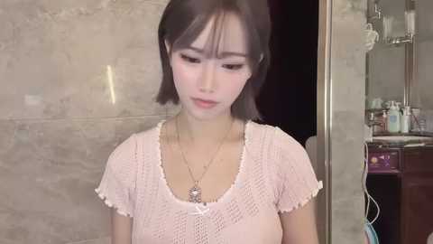 A video of an Asian woman with short, straight black hair, wearing a light pink crocheted top, standing in a bathroom with marble walls and a shelf of toiletries in the background.