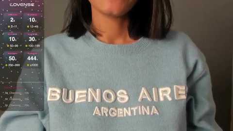 Video of a woman with medium skin tone, wearing a light blue Bu\u00f1os Aire Argentina sweatshirt, with a background featuring a weather forecast screen displaying temperature and precipitation.