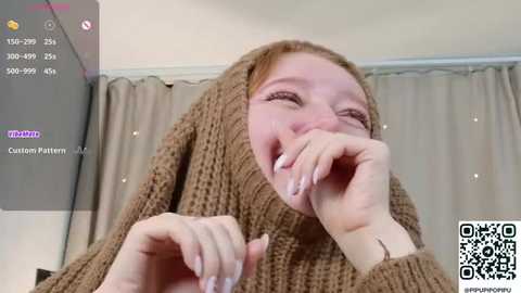 Video of a smiling woman with fair skin, wearing a textured, brown knit sweater, holding her hand to her mouth. Background features light-colored curtains. QR code in the bottom right corner.