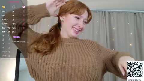 Video of a smiling, fair-skinned redhead with long hair, wearing a beige knit sweater, dancing with arms raised, against a beige curtain backdrop, with a QR code in the bottom right corner.