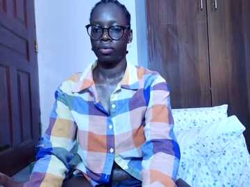 Video of a dark-skinned woman with short braided hair, wearing glasses and a plaid shirt, sitting on a bed with a floral pillow, in a simple bedroom setting.