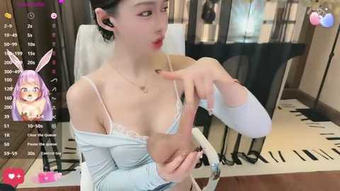 A video of a young East Asian woman with fair skin, wearing a light blue off-shoulder top and holding a baby. The background shows a modern room with a piano and a screen displaying a virtual avatar.
