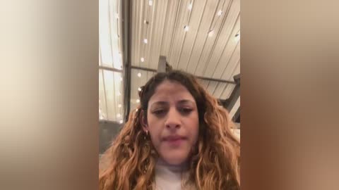 Video of a young woman with curly, shoulder-length hair, wearing a beige jacket, standing in a dimly lit indoor setting with a corrugated metal ceiling and string lights.