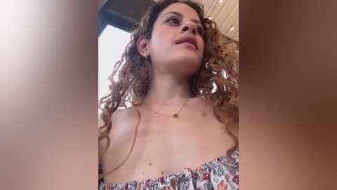 Video of a young woman with curly hair, wearing a floral off-shoulder top, standing in an indoor space with beige walls.