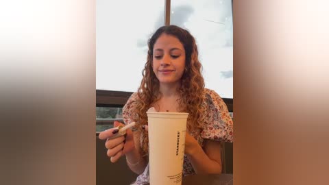 Video of a young woman with curly hair, wearing a floral dress, holding a white Starbucks cup and stirring it, seated indoors with large windows and blurred outdoor view.