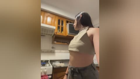 A video of a woman in a beige crop top and gray pants standing in a modern kitchen with wooden cabinets, white tiles, and a microwave.