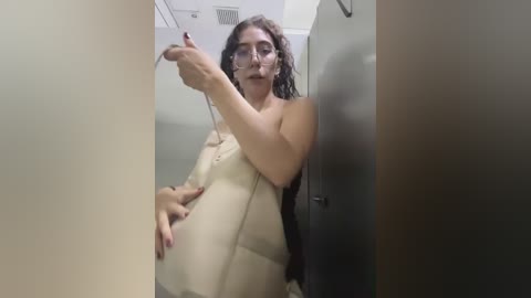 Video of a pregnant woman with long dark hair and glasses, wearing a beige coat, gesturing with her right hand, standing in a dimly lit, narrow hallway.