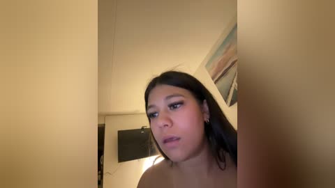 A video of a young woman with medium-length, straight black hair and light skin, wearing dark eyeliner and nude lipstick, standing in a dimly lit room with beige walls, a TV, and a framed picture in the background.