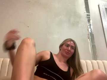 A video of a young woman with long blonde hair, fair skin, and medium build, wearing a black sports bra, sitting on a beige couch. The background features a textured white wall and a glass door.