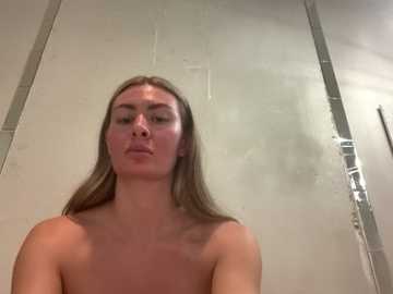A video of a topless young Caucasian woman with long, straight brown hair, fair skin, and small breasts, standing in a shower with a tiled glass door, water droplets on the glass.