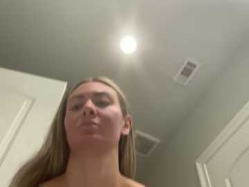 A video of a blonde woman with fair skin and closed eyes, taken from a low angle, showing her from the chest up. The background includes a ceiling light, white walls, and a door.