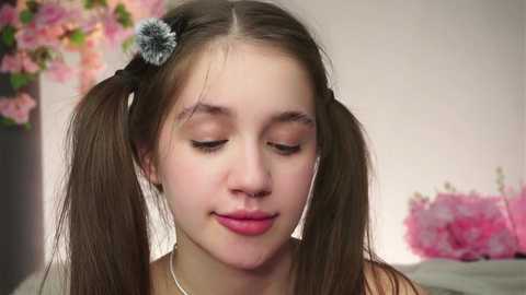 Video of a young, fair-skinned girl with pigtails and a gray scrunchie, wearing a light top, smiling softly, in a room with pink flowers and a soft-focus background.