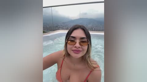 Video of a young woman with light brown skin, shoulder-length straight hair, and large sunglasses, smiling while standing in a hot tub with a cityscape and mountain view in the background.