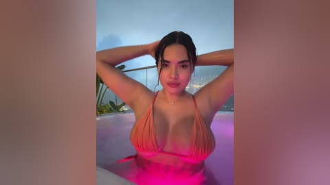 Video of a young, light-skinned woman with dark hair, wearing a coral bikini, arms raised, in a pool at sunset.