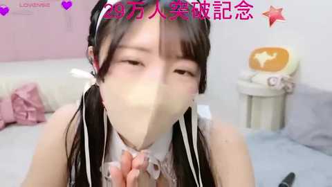 Video of an Asian woman with long black hair in pigtails, wearing a frilly maid outfit, holding a mask over her face, in a pastel-colored bedroom.