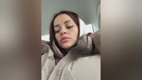 A video of a young woman with light skin, straight brown hair, and closed eyes, wearing a beige jacket and a light pink scarf. The background is blurred, suggesting an indoor setting.