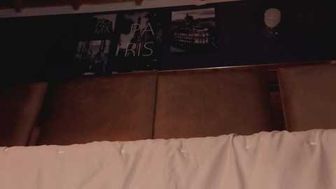 A dimly lit video of a bedroom with a wooden headboard, a white duvet, and a black wall-mounted clock. A poster of a black-and-white city scene with \"Paris\" text is partially visible above.