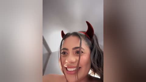 Video of a smiling woman with medium brown skin, wearing red devil horns and a playful expression. She has dark hair and is partially obscured by a white wall.