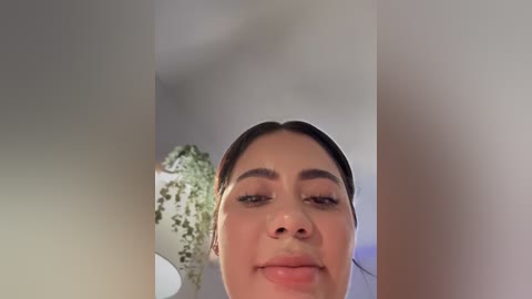 Video of a young woman with medium skin tone, dark hair tied back, and light makeup, smiling slightly, standing in a white-walled room with green plants in the background.