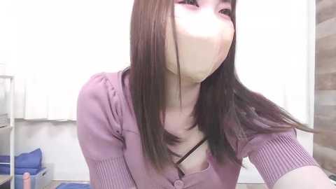 Video of a young Asian woman with long, straight black hair, wearing a purple cardigan, face covered with a white surgical mask, indoors with white walls and medical equipment visible in the background.