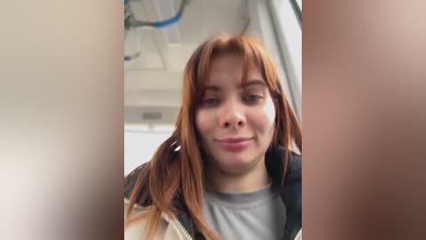 Video of a young woman with long, straight, auburn hair and fair skin, wearing a gray shirt and a plaid jacket, smiling slightly. Background shows hospital equipment.