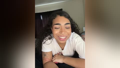 Video of a smiling, light-skinned Black woman with curly hair, wearing a white T-shirt, lying on a bed.