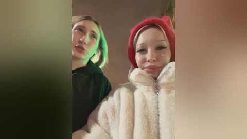 Video of two women, one with blonde hair in a black hoodie, and the other with a red hat and a white fluffy jacket, standing closely together.