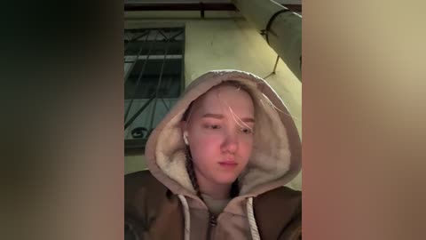 A video of a young woman with light skin, wearing a hooded jacket, sitting in a dimly lit room with a window and metal bars in the background.