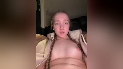 A video shows a young, fair-skinned, blonde woman with small breasts, lying on her back, partially undressed, wearing a partially open jacket, in a dimly lit room.