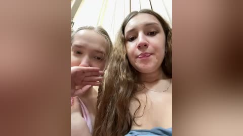 Video of two teenage girls with light skin, one with long brown hair and a light blue top, the other with light pink hair in a ponytail. They are smiling and close together, partially obscured by the image frame.