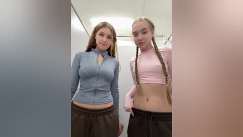 Video of two young women in a bathroom, one with long hair in a pink crop top, the other in a light blue zip-up, both in brown pants, standing close together.
