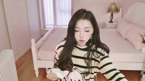 Video of a young Asian woman with long black hair, wearing a striped shirt, playing a video game in a pink-themed bedroom with a white bed and lamp.
