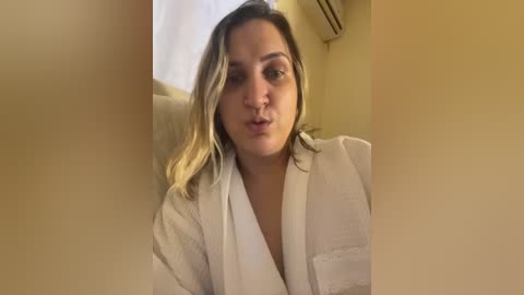 Video of a Caucasian woman with blonde hair, wearing a white robe, seated in a dimly lit room with beige walls and a window. She has a neutral expression and is slightly out of focus.