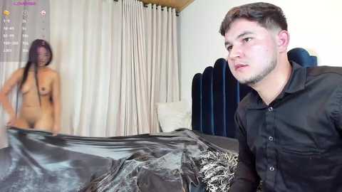 A video featuring a topless woman with dark skin and long braids standing naked beside a man in a black shirt, both in a bedroom with a bed covered in shiny, black plastic.