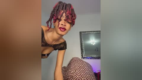 Video of a woman with short, dyed red and black dreadlocks, wearing a black lace bra, leaning over a bed with geometric-patterned sheets. The background features a framed mirror and a light switch.