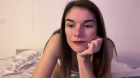 Video of a young woman with straight brown hair, fair skin, and medium build, lying on a bed with white sheets, resting her chin on her hand, wearing a white tank top, in a dimly lit room.