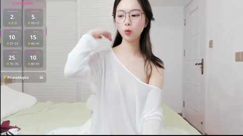 Video of an East Asian woman with straight, shoulder-length black hair, wearing glasses, a white off-shoulder sweater, and sitting on a bed in a white bedroom.