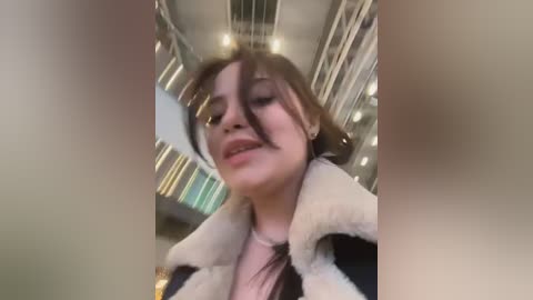 Video of a young woman with light skin, brown hair, and a white fur coat, standing in a modern indoor setting with a blurred background.